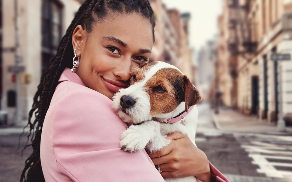 Mujer sosteniendo un perro con un Collar de Mascota de Pandora Moments. Mujer llevando pendientes de aro con charms de color rosa.
