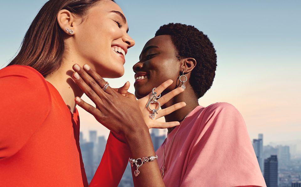 Due donne che indossano orecchini e anelli Pandora, basi per charm, portachiavi e bracciali Pandora Moments con charm a cuore