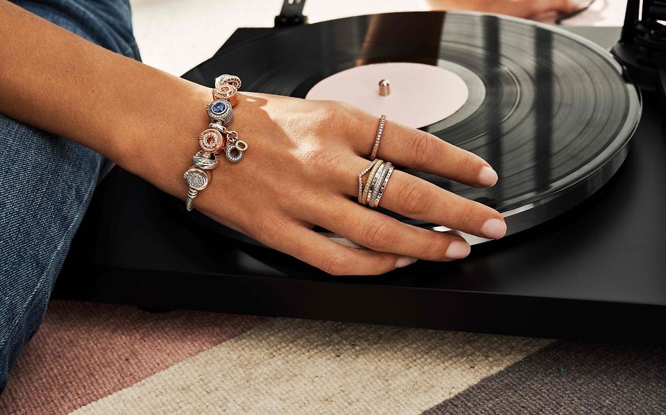 一只纤纤玉手置于黑胶唱片上，手指上叠戴着Pandora新经典系列戒指，腕上戴有一条串饰手链。